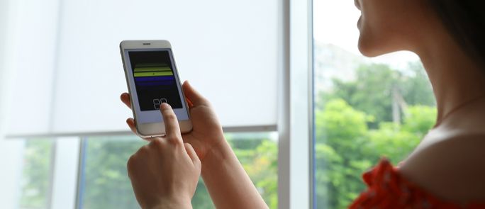 Woman Standing Near Window Operating A Motorized Shade With A Remote.